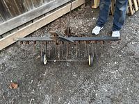 Rusty metal frame lawn irrigator with multiple spike tines, two wheels, and long handle, on gravel surface