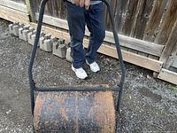Side view of lawn roller with person's legs visible, showing rusted metal drum and black metal handle.