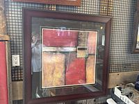 Framed modern abstract art with geometric shapes and a warm color palette. Shows the full front view of the artwork and frame on a wall.