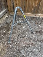 Full view of the steel satellite dish tri-pod stand placed on gravel ground against a wooden fence.