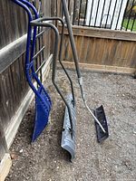 Three snow removal tools standing against a wooden fence in outdoor setting, showing overall shape and handle design.