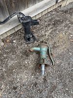 Two cast iron antique water pumps on dirt ground next to a wooden fence, one black and one greenish patina.