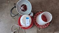 Two plastic chicken feeders with red bases and white cones, hanging cords visible, along with silver reflective heat lamp with red bulb and black power cord seen from above