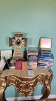 Cream-colored decorative cross with heart cutout and Bible verse alongside a stack of religious books and CDs on a decorative gold console table against a teal wall.