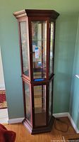 Full view of the tall corner display cabinet showing angled glass and wood construction with dark brown finish in a corner setting.