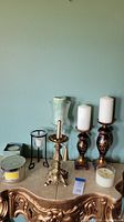 Full view of all items on decorative surface, including ornate gold-tone candlestick holders with pillar candles, glass candle holders, extinguishing snuffer, and various jar candles.