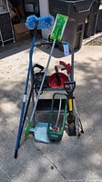 Photo showing assortment of household utility items arranged together on pavement including two blue fluffy dusters, dust pans in red, black and light blue, a green garden grabber, a wooden cane, a hand cultivator tool, and small step stools.