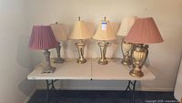 Full view of six assorted decorative table lamps on a white folding table against a wall.