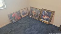 Four framed prints leaning against wall on dark blue carpet, showing various floral and thematic designs.