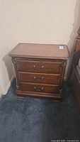 Front view of wooden nightstand showing three drawers and decorative corner carvings. Dark wood finish with wear marks.