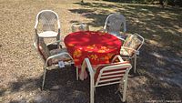 Photo showing round glass top table with 4 white metal chairs having floral cushions and a red floral tablecloth outdoors on ground.