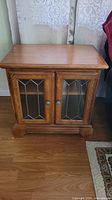 Front view of one wooden end table showing glass panel doors and brown finish.