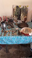 Full view of all items including silverplate tea set, pitcher, candelabra, flatware, swan figurines, large tray, and decorative wall hangings on blue floral tablecloth.
