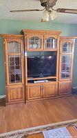 Full frontal view of oak entertainment center with 3 main sections, leaded glass doors on upper side and glass cabinets sides, space for TV and electronic devices in center lower section.