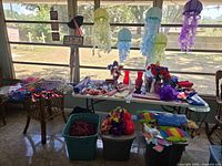 Wide view of table with assorted colorful summer party decorations including paper lantern jellyfish, tiki torches, floral leis, hula skirts, and 4th of July-themed decorations including flags and pinwheels.
