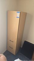 Left angled view of a tall office cabinet in light wood with two handles in a corner of a room.