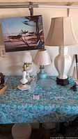Three vintage table lamps of varying sizes on a blue floral patterned tablecloth along with clear glassware and decorative wall hanging.
