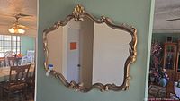 Front view of large vintage wall mirror mounted on wall showing overall ornate gold tone gilded frame with decorative scrolls and flourishes.