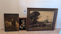 Three wall art pieces: raccoon painting, classical-style Aristotle painting, and framed landscape painting; all shown upright against a wall on floor tiles.