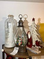 Large glass light-up bottle with white glittery tree inside and cork top, next to water-filled glass lantern with snowman figure inside and light, wooden Christmas tree with red star topper and decorations including Santa figure and red cardinal.