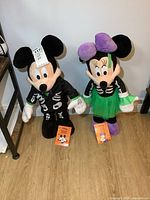 Front view of both Mickey and Minnie Mouse Halloween greeters standing side by side on a wooden floor against a white wall, showing their full costumes and size.