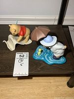 Two Disney Winnie the Pooh figures displayed on a shelf, one Pooh with umbrella and blue puddle base with yellow duck, other Pooh kneeling with red shirt reading scroll.