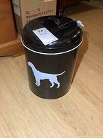 Full view of the black enamelled metal pet food can on a wooden floor, showing white dog silhouette design.