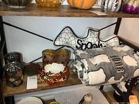 Shelf with Halloween-themed throw blanket with glow in the dark eyes and 'BOO!' text, ghost pillow with 'Spooky' text, pumpkin candle holder, and other fall decor.