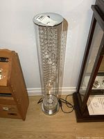 Full view of the tall cylindrical LED floor chandelier with hanging clear crystal bead strands inside the glass tube, metal base and top caps visible.