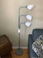 Full view of the 3-headed metal floor lamp with white plastic shades against a wall near a gray couch.