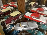 Three diecast cars displayed on glass shelf: silver Mercedes-Benz 300SL coupe, maroon and beige vintage convertible, red and white classic car.