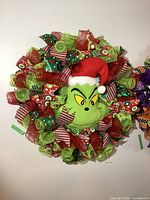 Front view of round Christmas wreath with plush Grinch center, decorated with colorful mesh and ribbons in green, red, and festive patterns.