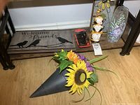 Wooden Welcome Friends sign, screwdriver set, faux flower arrangement in black metal wall vase, part of decorative glass vase with floral design, and resin figurine visible on shelf.