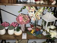 Shelf with three small white planters holding faux succulents, two bowls with pink faux flowers, tall vase with faux orchid stems, white metal lantern with faux greenery, and resin gnome figurine