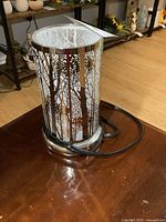 Full view of the lamp showing the cylindrical shape and forest tree design cut into metal, polished metal base, and attached electrical cord.