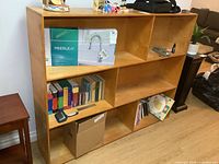 Front angled view of wooden bookshelf showing contents in some compartments including books, boxed faucet, and small items.