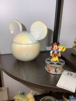 Porcelain Mickey ears candle container with iridescent glaze next to a plastic Mickey Mouse in director's chair on a film reel base with character images.