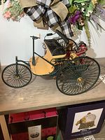 Left-front angle view of the Franklin Mint 1886 Benz Motorwagen diecast model showing front single spoked wheel, green and beige frame, black seat, and brass engine parts.