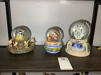 Three Disney musical globes displayed side by side: Snow White wind up globe, Winnie the Pooh stationary globe, and Cinderella battery-operated globe with castle and carousel.
