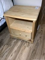 Front view of one unfinished solid pine night stand, showing single drawer with knob and open shelf above it, sitting on wood floor.