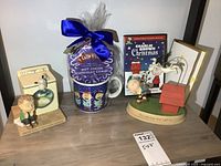 Full lot shot showing two Hallmark Peanuts figurines, large hot cocoa mug with blue bow and Cafe Du Monde hot cocoa mix, Charlie Brown Christmas DVD and card on table.
