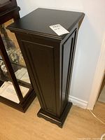 Black wooden pedestal plant stand with paneled sides and flat removable top, standing on wood floor near a wall and glass cabinet.