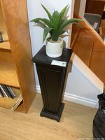 Full view of black plant stand with faux plant on top against wall, next to bookshelf and baseboard.