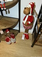 Photo of two plush figures on floor near shelving, showing larger figure standing and smaller weighted doorstop figure seated.