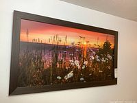 Framed printed artwork showing a scenic sunset over wildflowers and tall grasses, hung on a white wall.