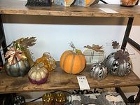 Shelf display of eight decorative pumpkins including glass, resin, metal, and fabric materials in various colors, shapes, and sizes as seasonal decor items.