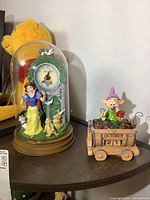 Snow White dome clock with woodland animals inside glass dome with wooden base, alongside dwarf train car perpetual calendar with Dopey figure