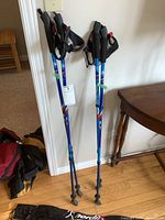 Two pairs of blue Nordix walking poles with black ergonomic handles and wrist straps leaning against a wall next to a brown wooden table and some backpacks on the floor.