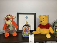 Tigger plush holding a globe clock, framed Winnie the Pooh print, music snow globe, and Winnie the Pooh plush on wooden shelf.