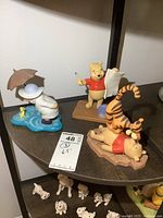 Shelf with three Winnie The Pooh-themed porcelain figurines: Tigger on Pooh, Pooh with artist paintbrush and paint bucket, Pooh in raincoat with umbrella and yellow duck.